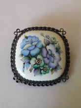 Charger l'image dans la galerie, Cette broche ancienne, ornée de fleurs sur un cabochon en porcelaine, est un joli modèle serti de métal argenté travaillé. Blanc, Bleu, Vert, Violet