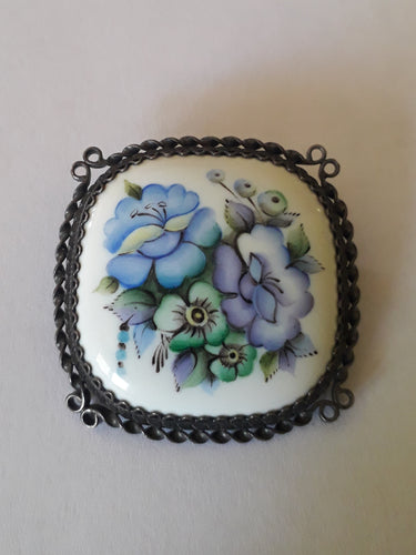 Cette broche ancienne, ornée de fleurs sur un cabochon en porcelaine, est un joli modèle serti de métal argenté travaillé. Blanc, Bleu, Vert, Violet